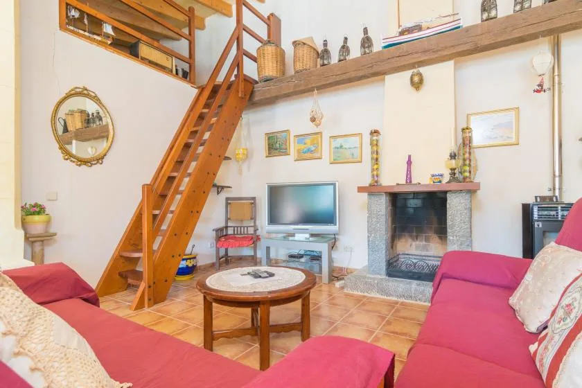 Una sala de estar con asientos, televisor y chimenea bajo una escalera de madera hacia un altillo.