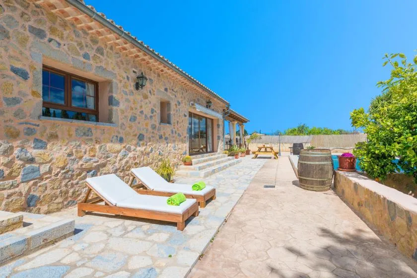 Una casa de piedra da a un patio soleado con tumbonas y un barril.