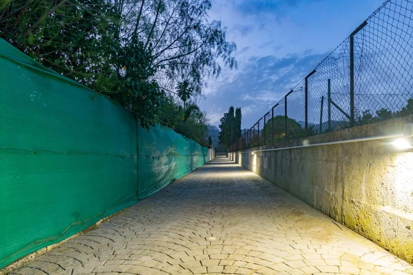 Un camino exterior pavimentado pasa entre una pantalla verde y un muro vallado.