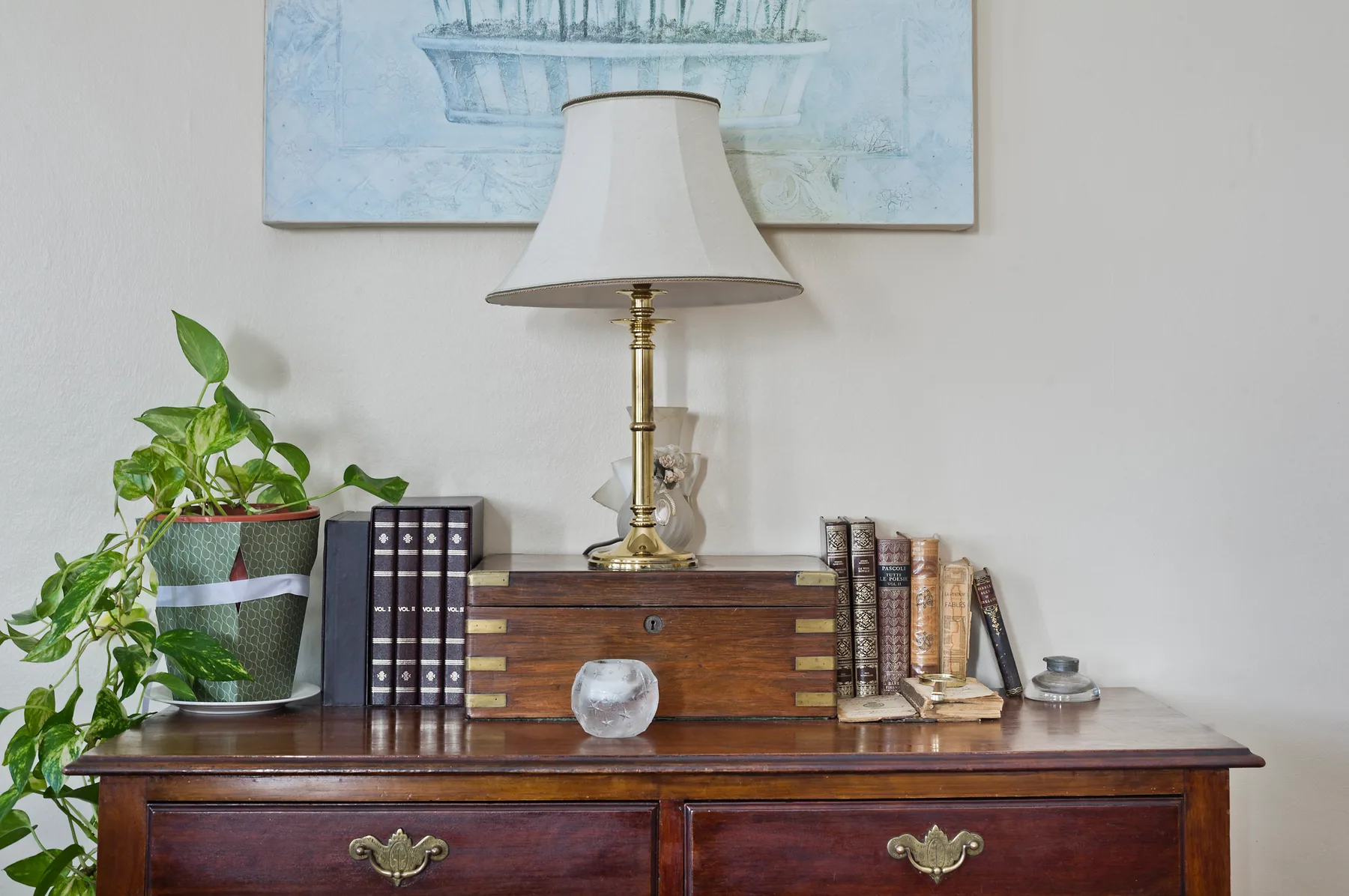 Una cómoda de madera con una lámpara, libros y una planta en maceta está junto a la pared.