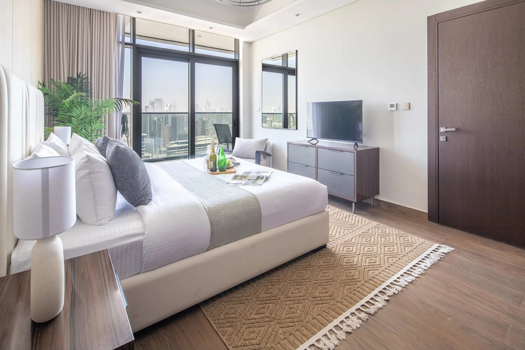 Un dormitorio luminoso con una cama, un televisor y grandes ventanas con vistas a la ciudad.