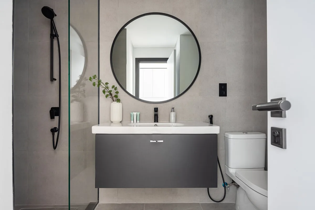 Un baño moderno con lavabo suspendido, espejo redondo y mampara de ducha de vidrio.