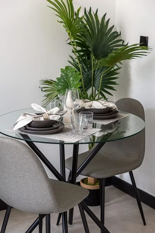 Una mesa de comedor está puesta con platos y cristalería junto a una planta frondosa.