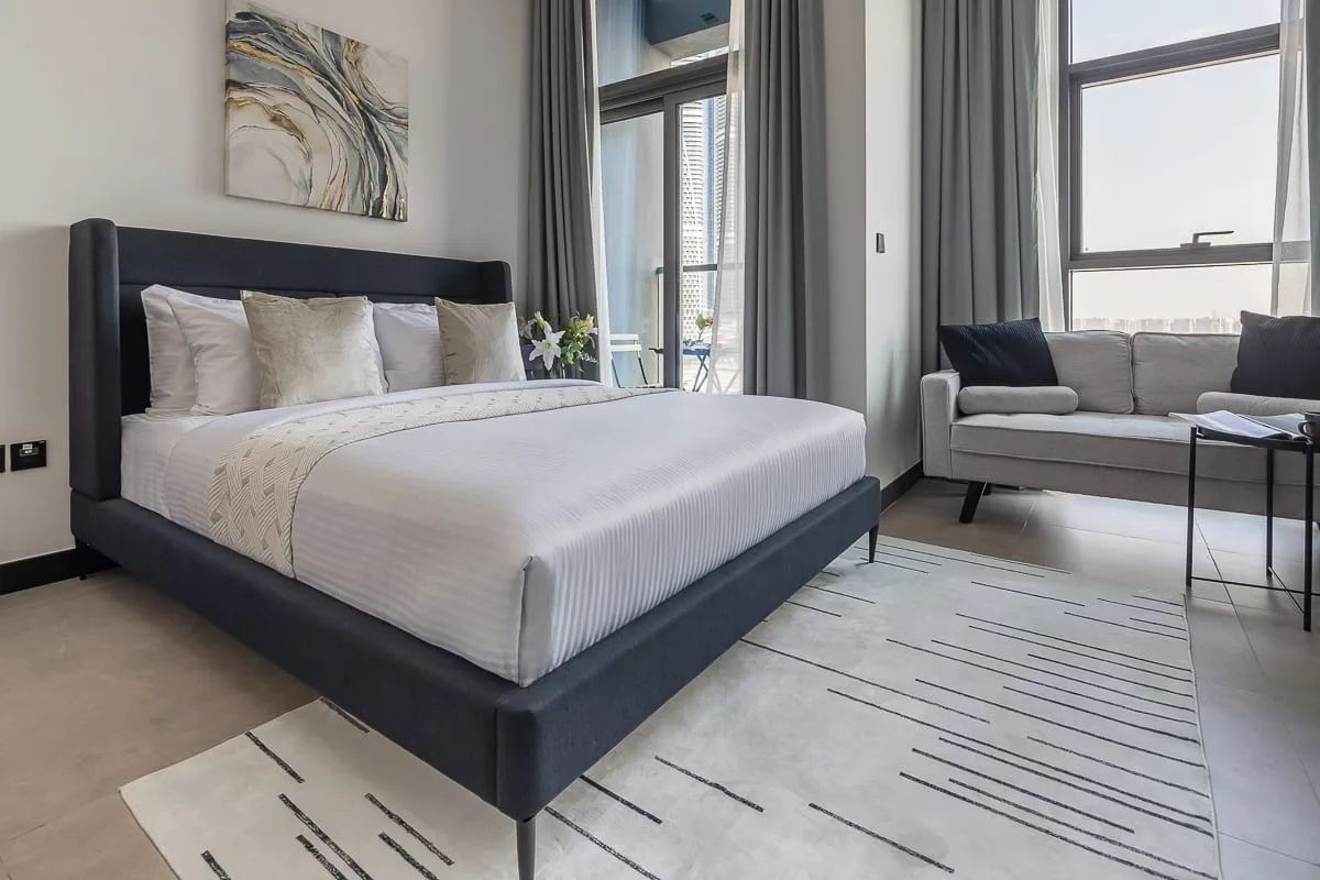 Un dormitorio moderno con una cama bien arreglada y una zona de estar junto a ventanas altas.