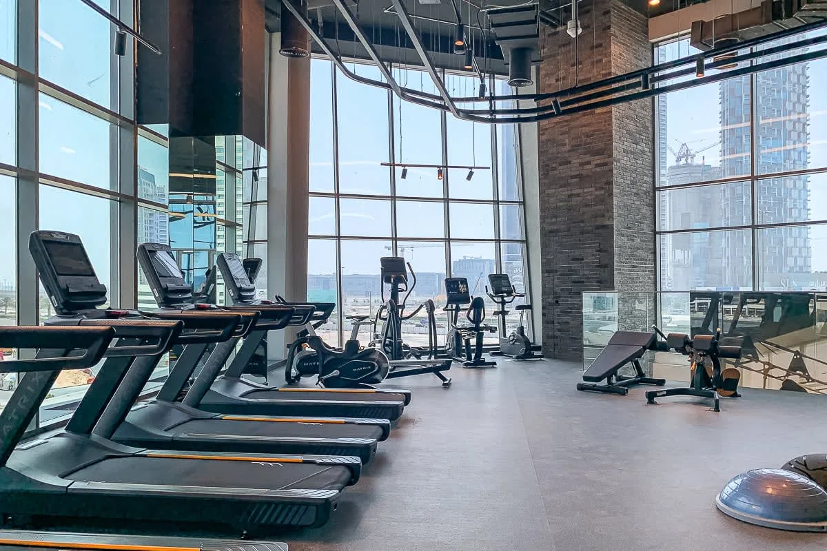 Un gimnasio con máquinas cardiovasculares y equipo de ejercicio junto a grandes ventanales.