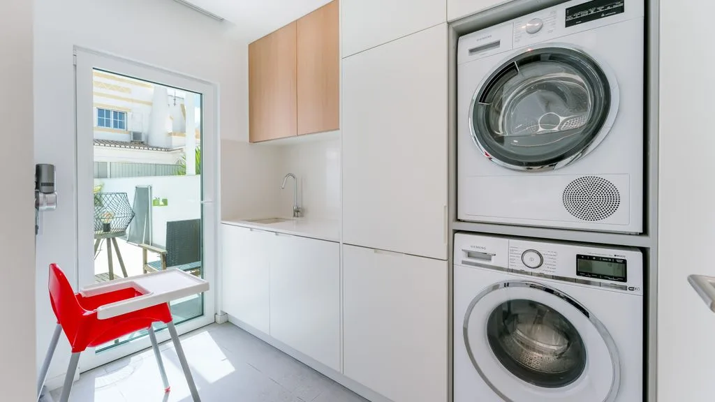 A bright laundry room with stacked laundry machines and a red chair by the glass door.