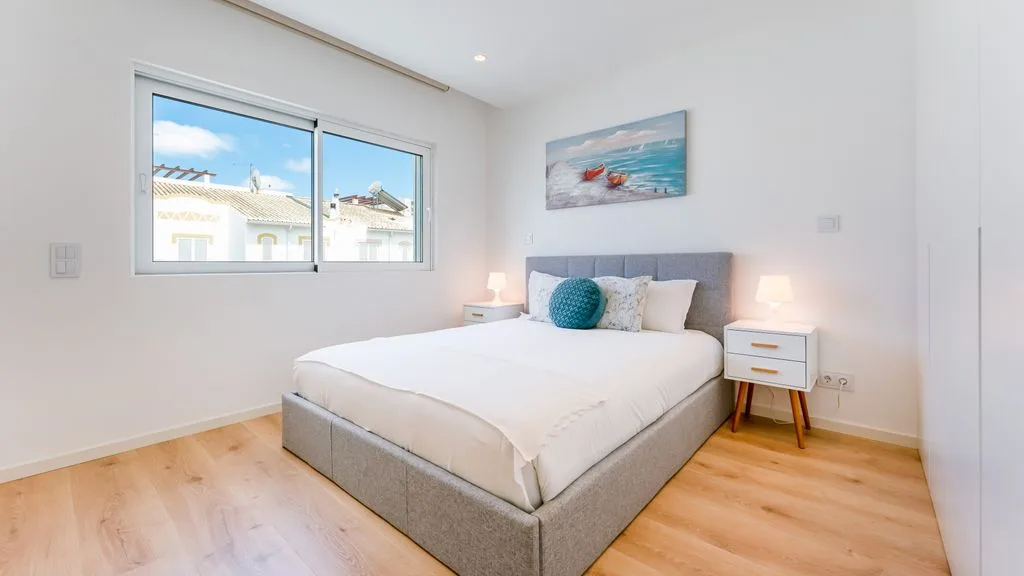 A bright bedroom with a bed, bedside tables, and a large window.