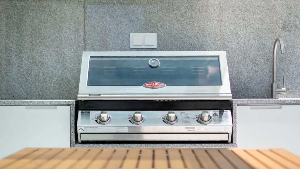An outdoor kitchen grill is set into a gray wall beside a wooden counter.