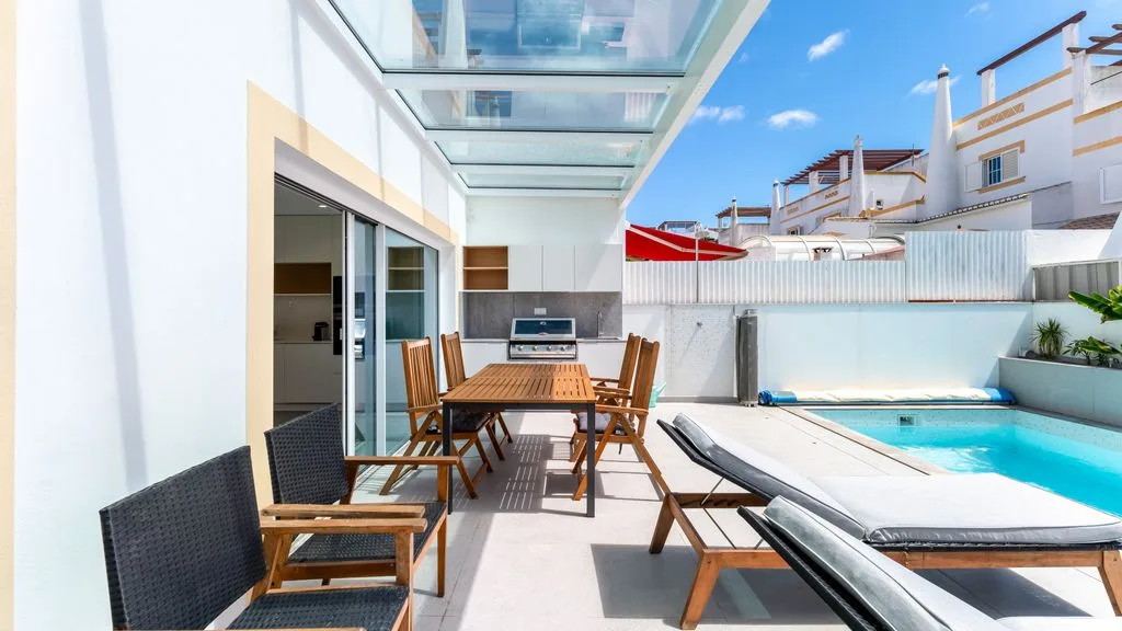 A sunny outdoor patio with a swimming pool and seating beneath a glass canopy.