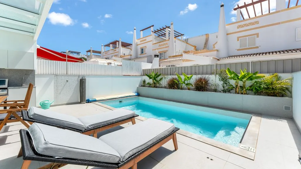 A sunlit pool patio with cushioned loungers and white buildings in the background.