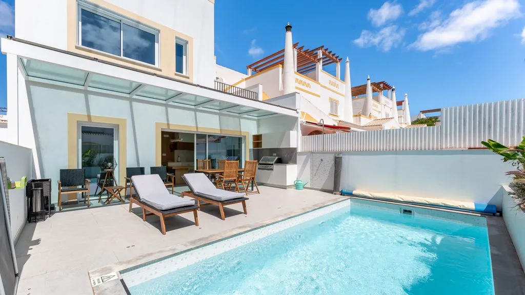 A sunny patio with a swimming pool and outdoor seating beside a modern house.