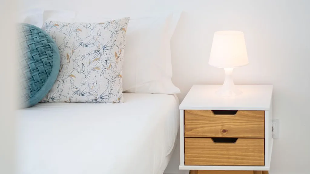 A bright bedroom with a white bed and a bedside table topped by a lamp.