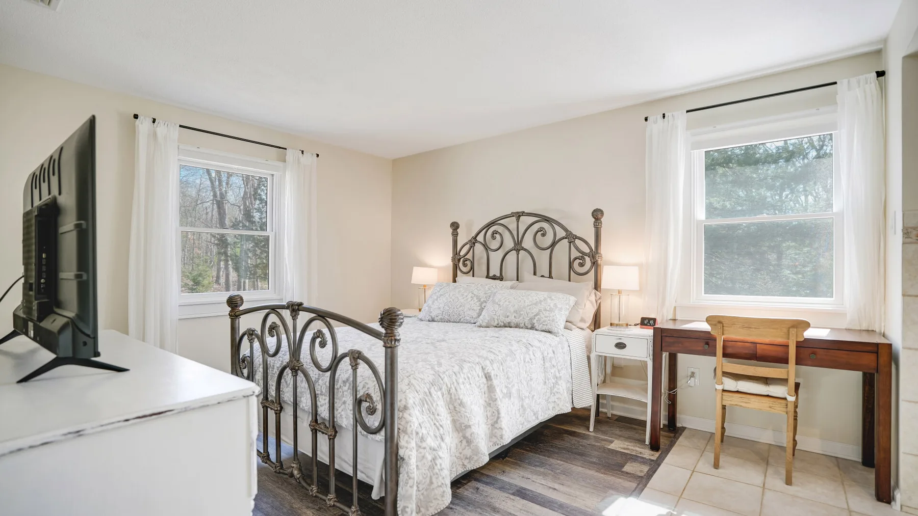 A bedroom with a metal bed, two windows, and a wooden desk.