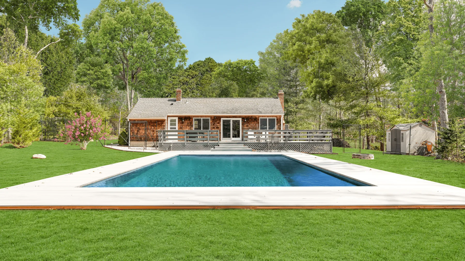 An outdoor swimming pool sits in front of a house, surrounded by lawn and trees.