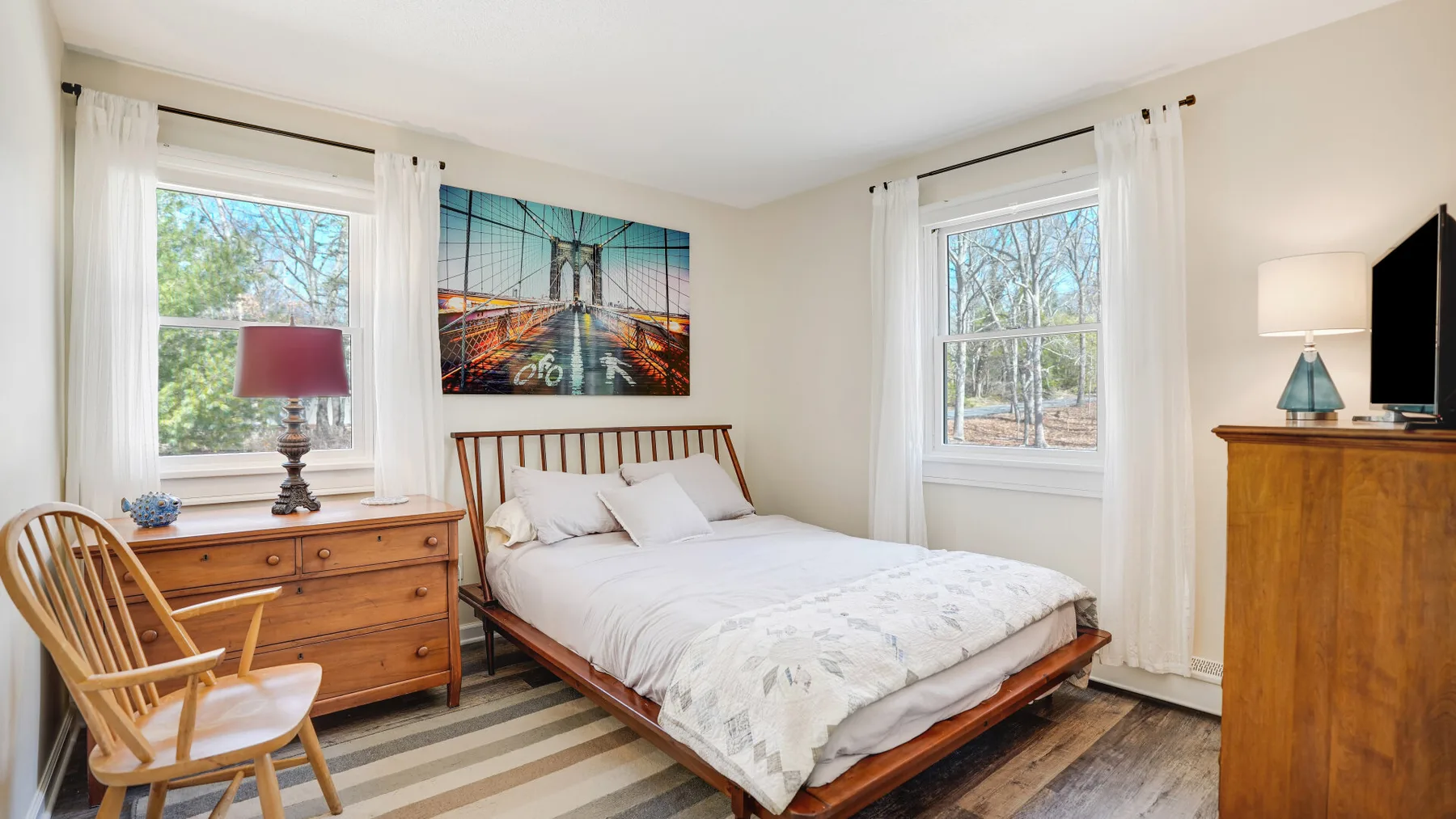 A bright bedroom with a bed, wooden dresser, and windows overlooking trees.