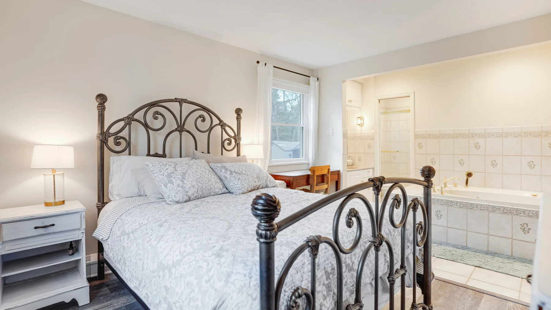 A bedroom with a bed beside a window and an adjoining tiled bathtub area.