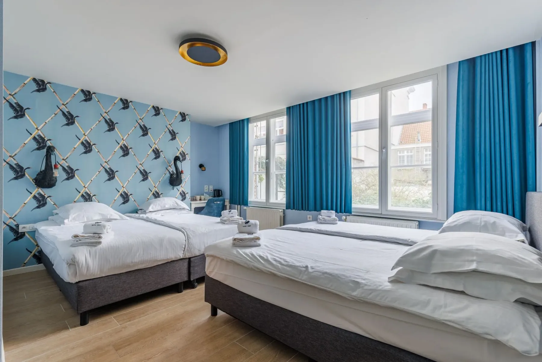 Habitación de hotel con dos camas individuales, papel pintado azul con motivos de cisnes y cortinas azules del suelo al techo.