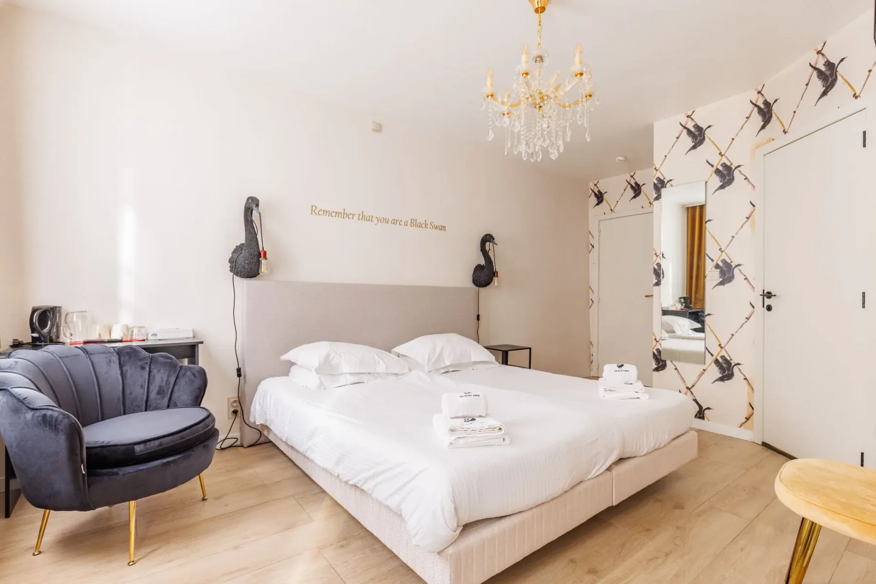 Habitación de hotel con una cama grande, un sillón gris, una lámpara de araña dorada y papel pintado con motivos de aves.