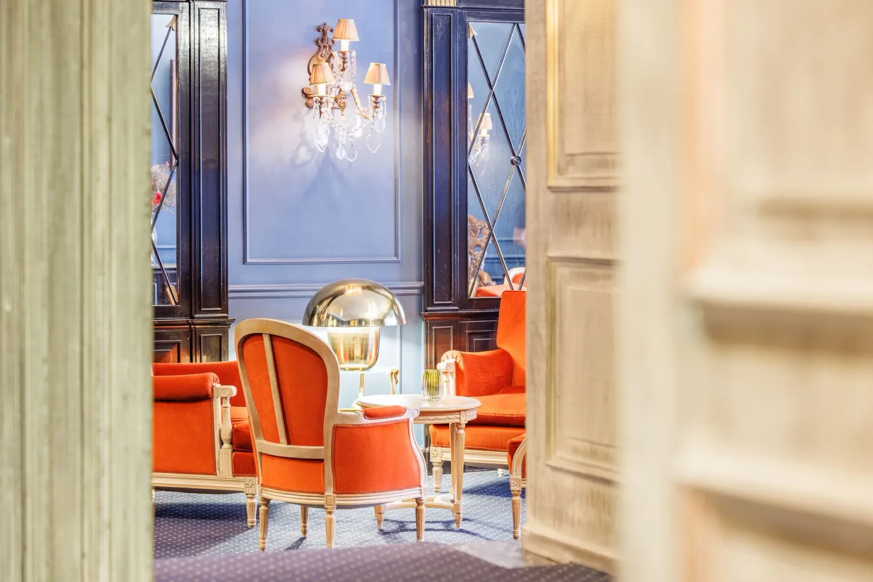 Orange armchairs and a small side table in a blue room with ornate mirrors and a wall sconce.