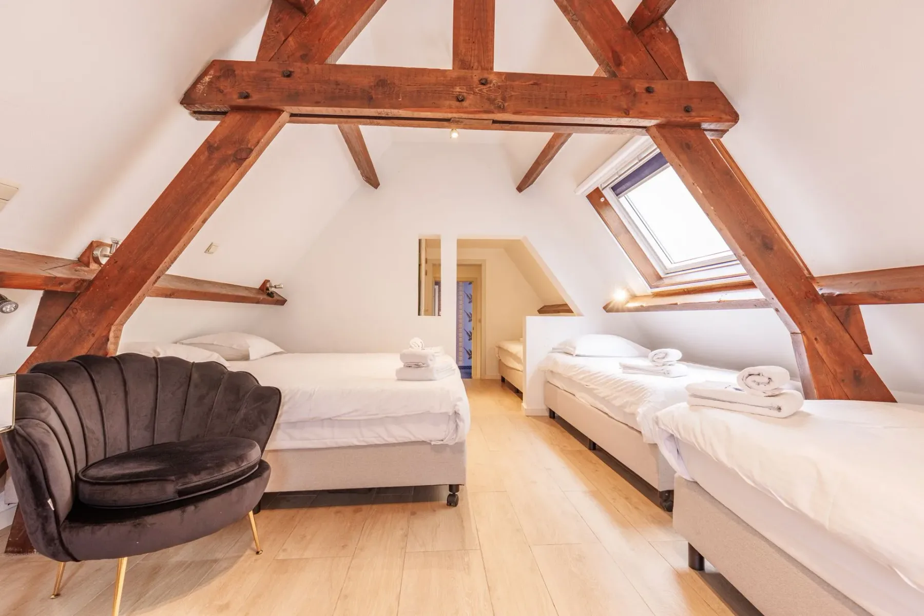 Dormitorio abuhardillado con vigas de madera vistas, una silla de terciopelo gris carbón y tres camas blancas bajo un techo inclinado con una claraboya.