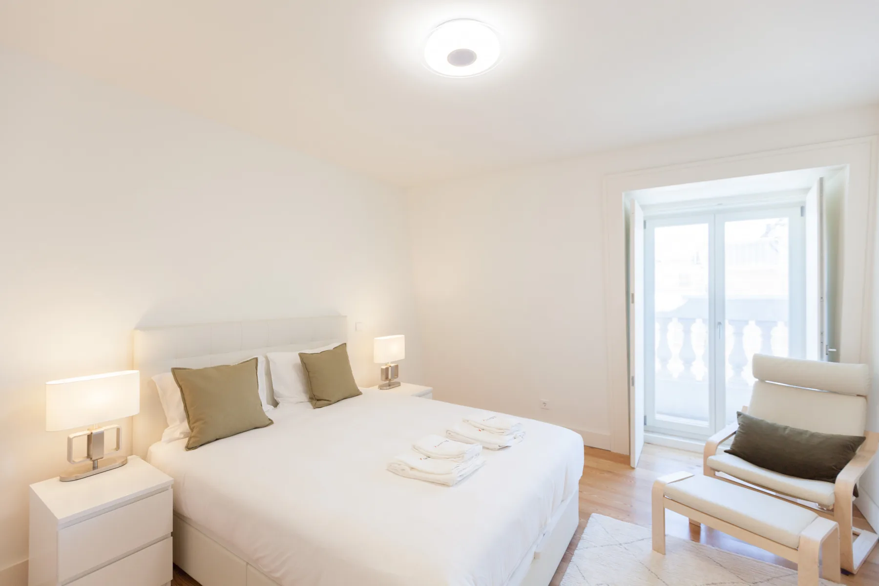 Un dormitorio luminoso con una cama bien hecha y una silla junto a las puertas del balcón.