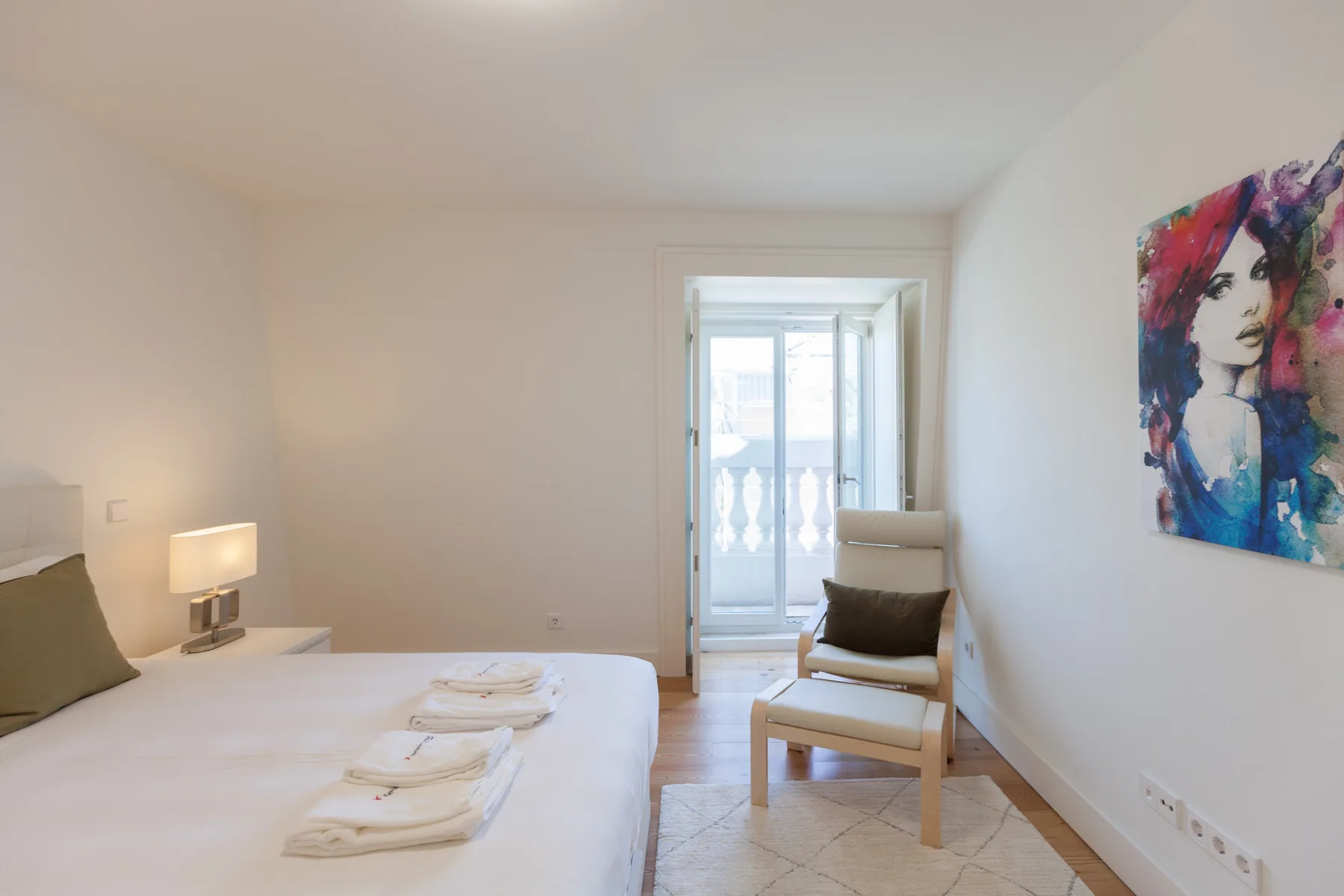 Dormitorio luminoso con una cama, un asiento y una obra colorida en la pared.