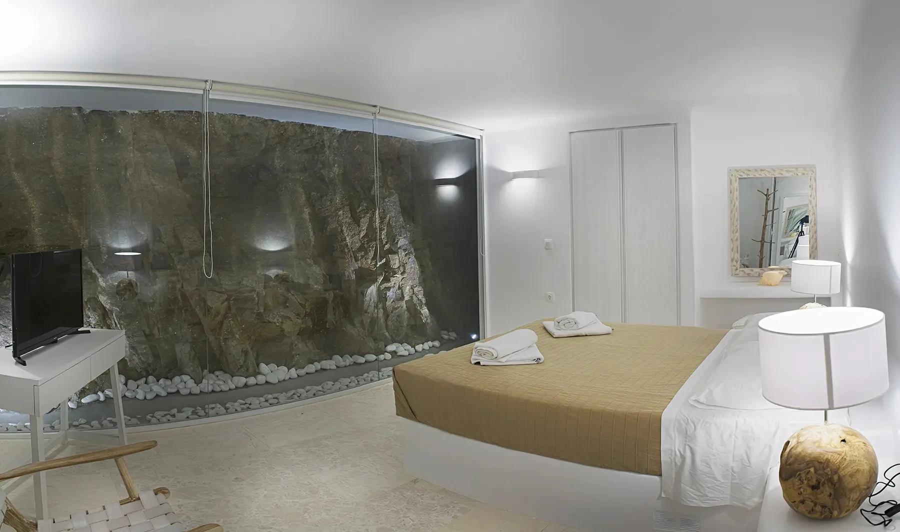 A bedroom with a bed beside a large rock feature behind glass.