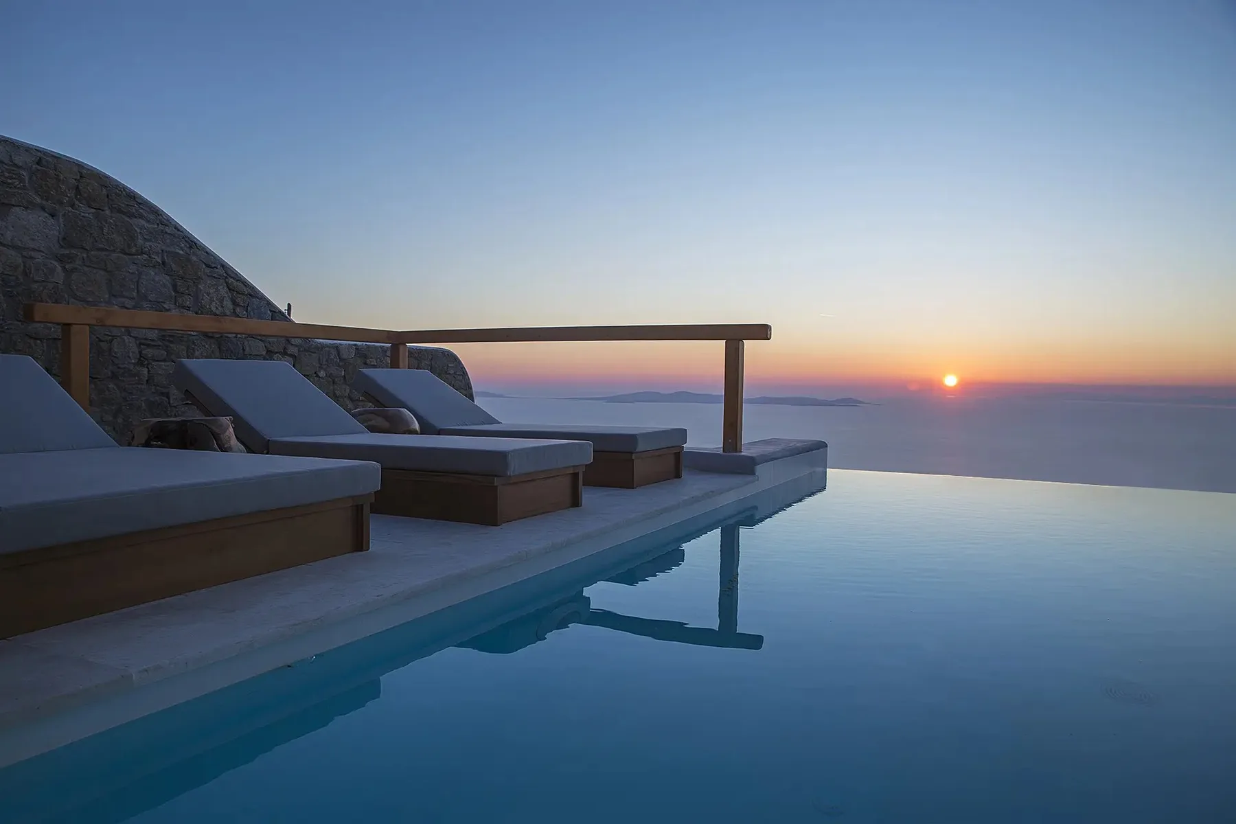 An infinity pool at sunset overlooks the sea, with loungers along the edge.
