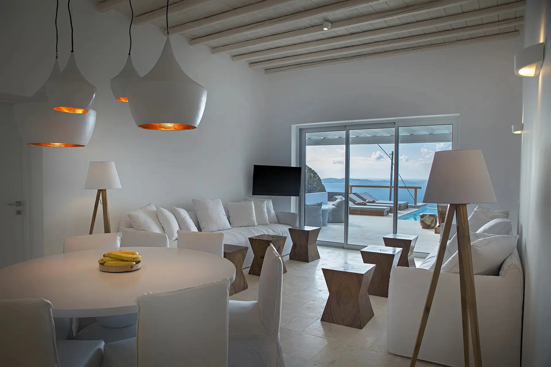 A bright living and dining area has white seating and large windows overlooking the sea.