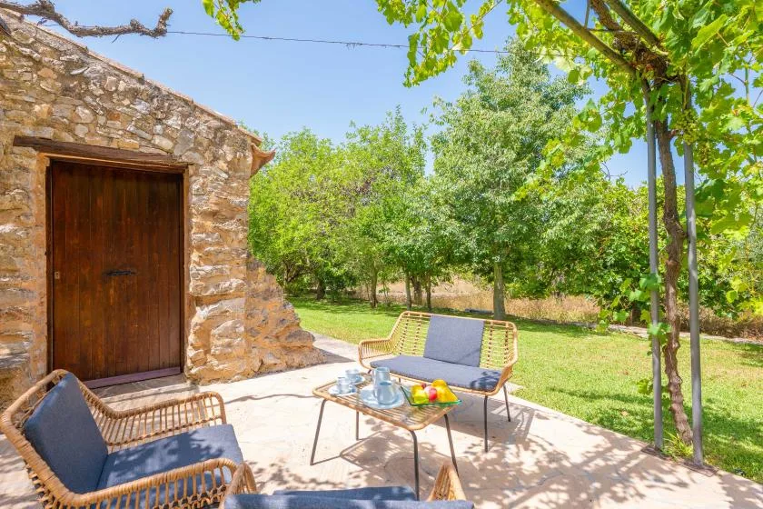 A stone building opens onto a shaded patio with seating and a garden.