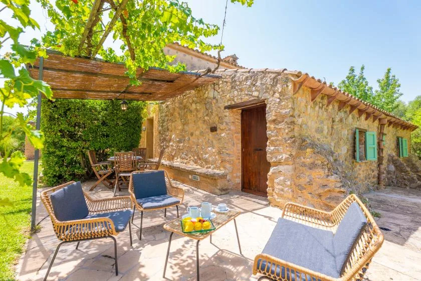 A sunny stone-house patio with outdoor seating beneath a leafy pergola.
