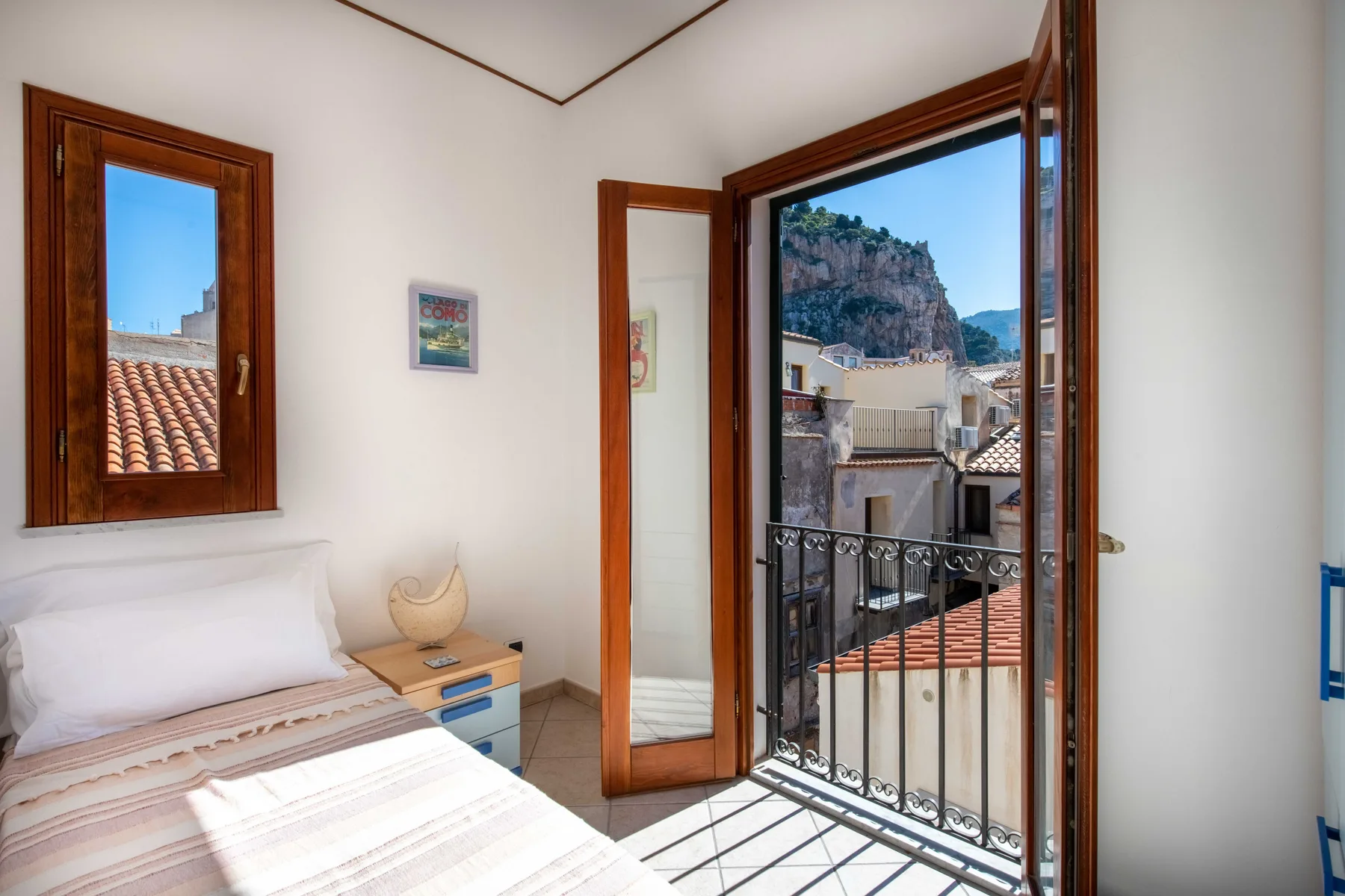 A bedroom with a bed beside an open balcony door overlooking nearby buildings.