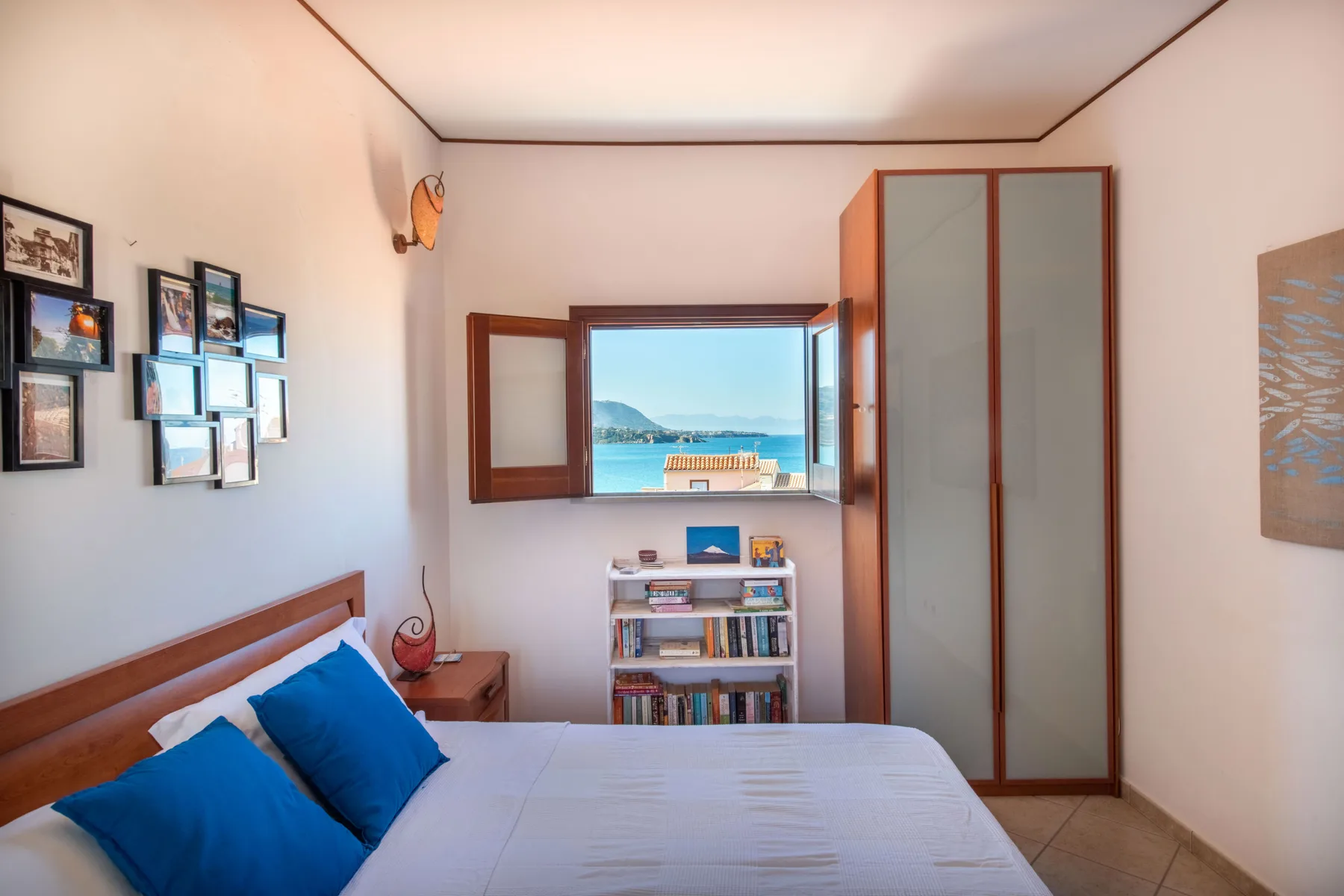 A bedroom with a bed, framed pictures, and a window overlooking the sea.