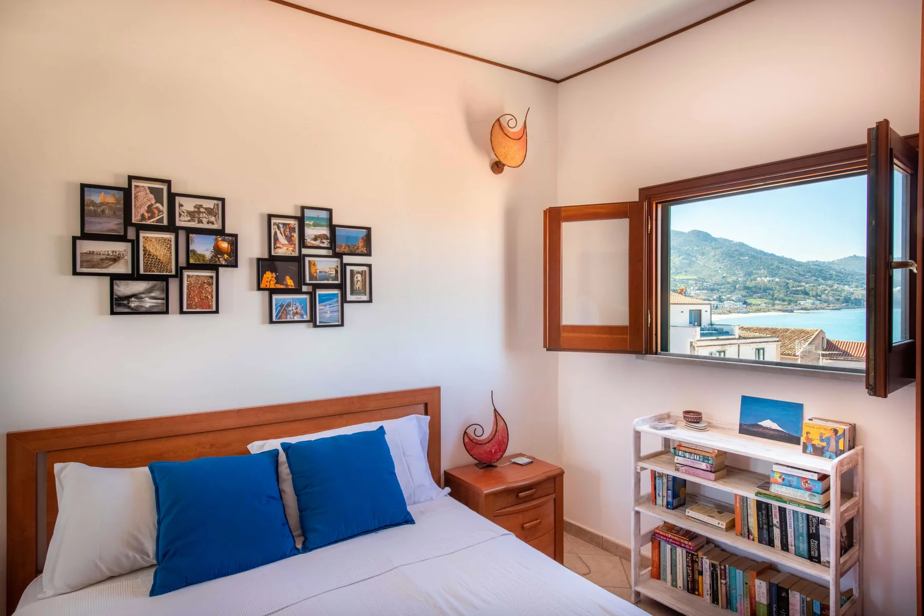 A bedroom with a bed, framed pictures, and an open window overlooking the water.