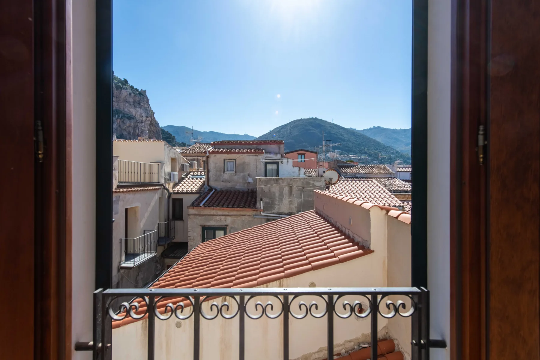 A mountain view overlooks terracotta rooftops from a window with a metal railing.