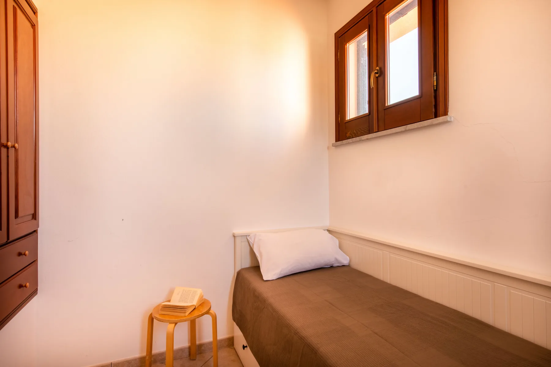 A small sleeping area with a single bed, a stool, and a window.