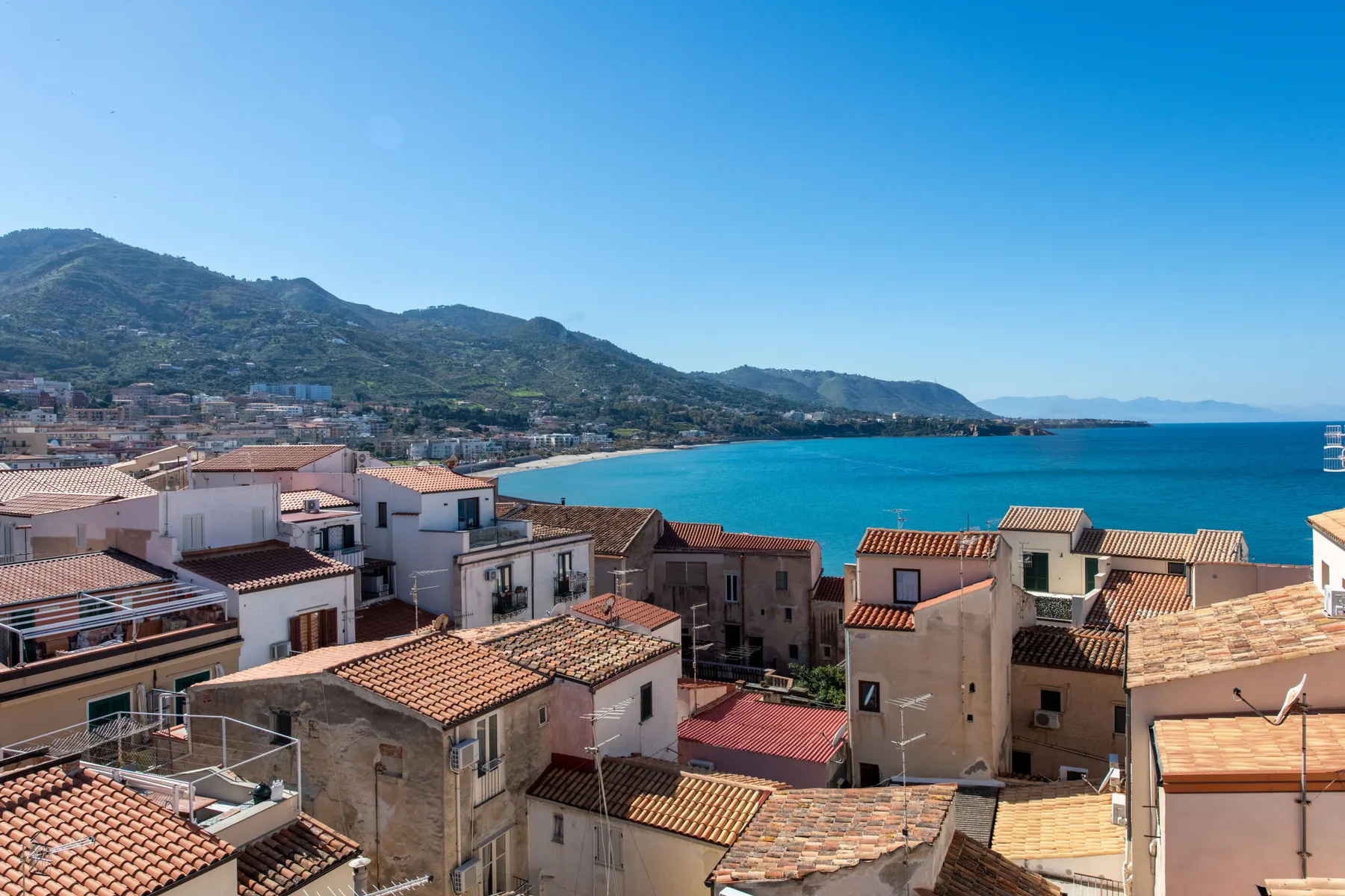 A coastal town with terracotta rooftops overlooks the blue sea and surrounding hills.
