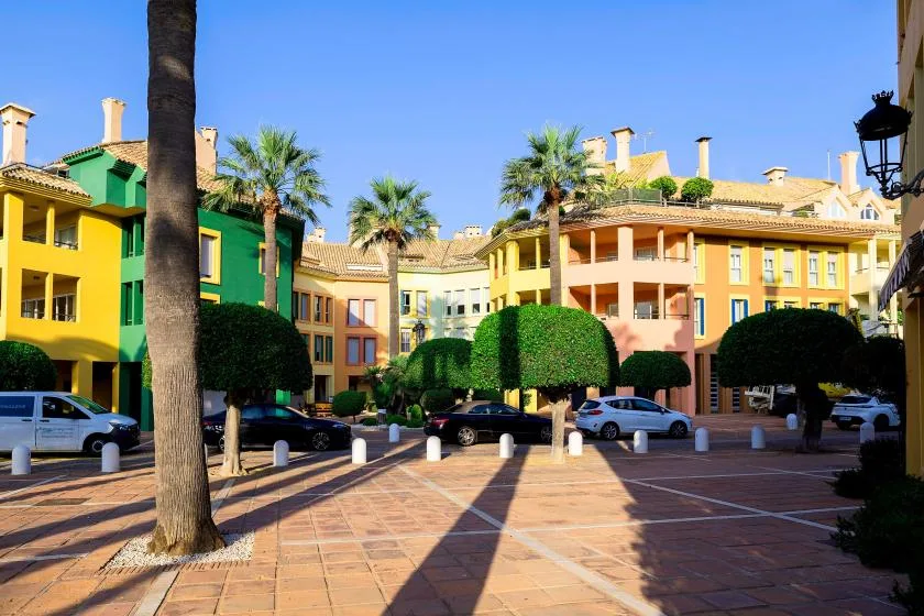 Un patio soleado pavimentado, rodeado de edificios coloridos, palmeras y vegetación recortada.