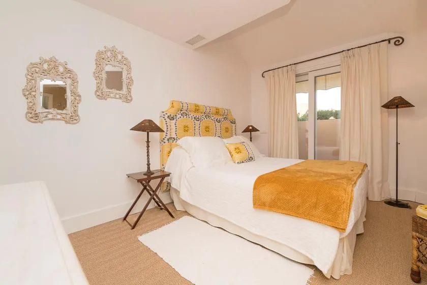 Un dormitorio luminoso con una cama blanca, cojines estampados y cortinas junto a la ventana.