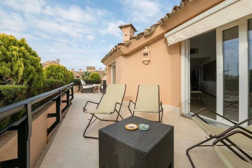 Una terraza soleada con asientos reclinables y una mesa pequeña junto a puertas corredizas de vidrio.