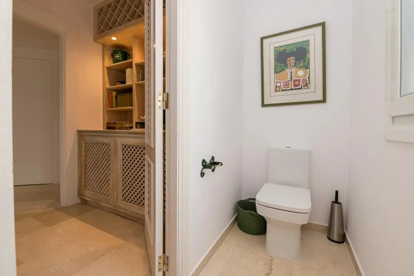 Un baño con un inodoro, una obra enmarcada y una cesta tejida junto a la puerta.