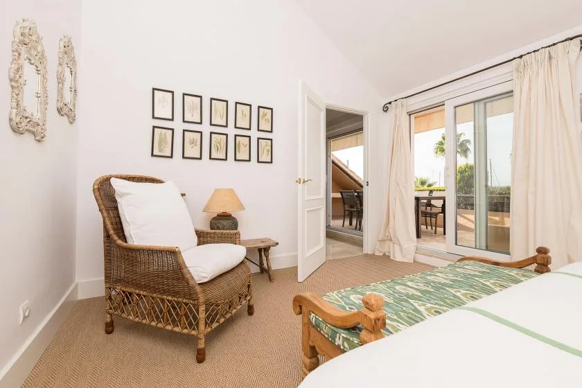 Un dormitorio con un sillón de mimbre junto a una cama y puertas de cristal hacia un balcón.