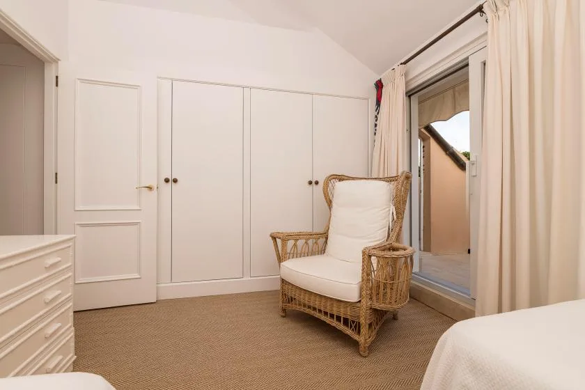 Una zona de estar en un dormitorio, con un sillón de mimbre junto a una puerta de vidrio con cortinas.