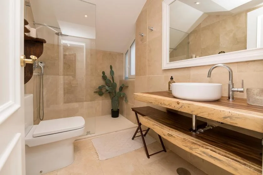 Un baño con azulejos beige, ducha de vidrio, lavabo sobre encimera y una planta.