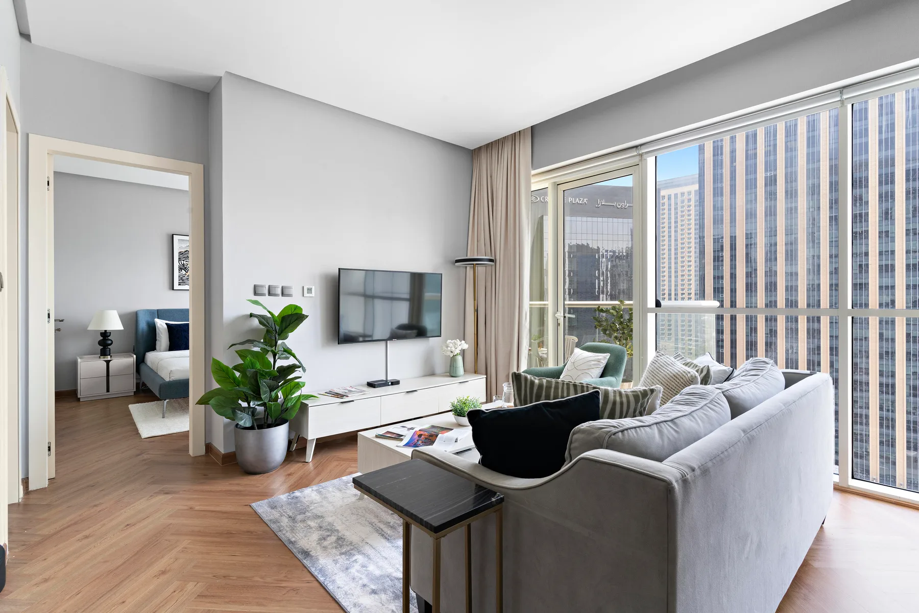 A bright living room with seating, a television, and large windows overlooking nearby buildings.