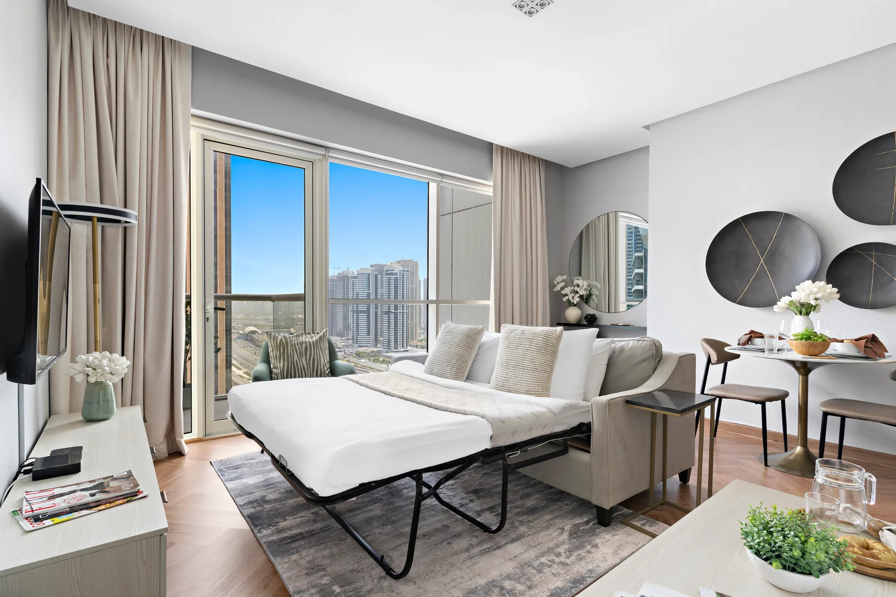 A bright bedroom with a bed, seating area, and large windows overlooking city buildings.