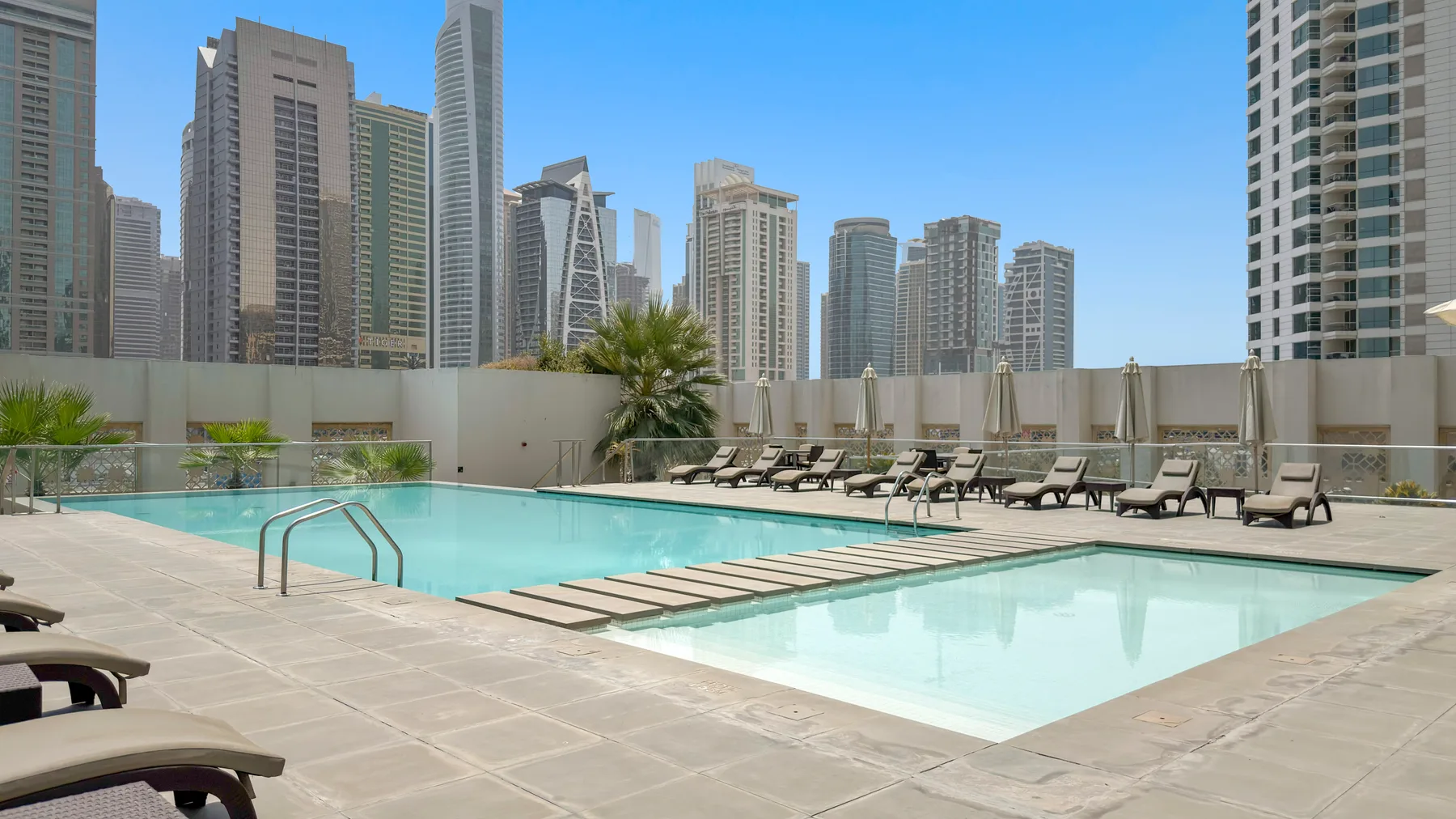 An outdoor swimming pool and shallow pool sit among lounge chairs, with high-rise buildings in the background.