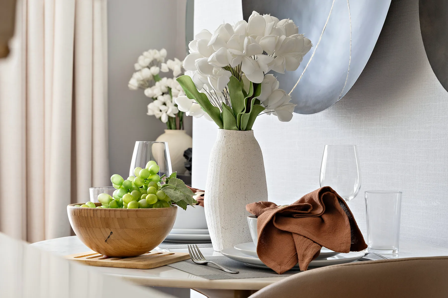 A dining table holds a vase of white flowers, green grapes, and tableware.