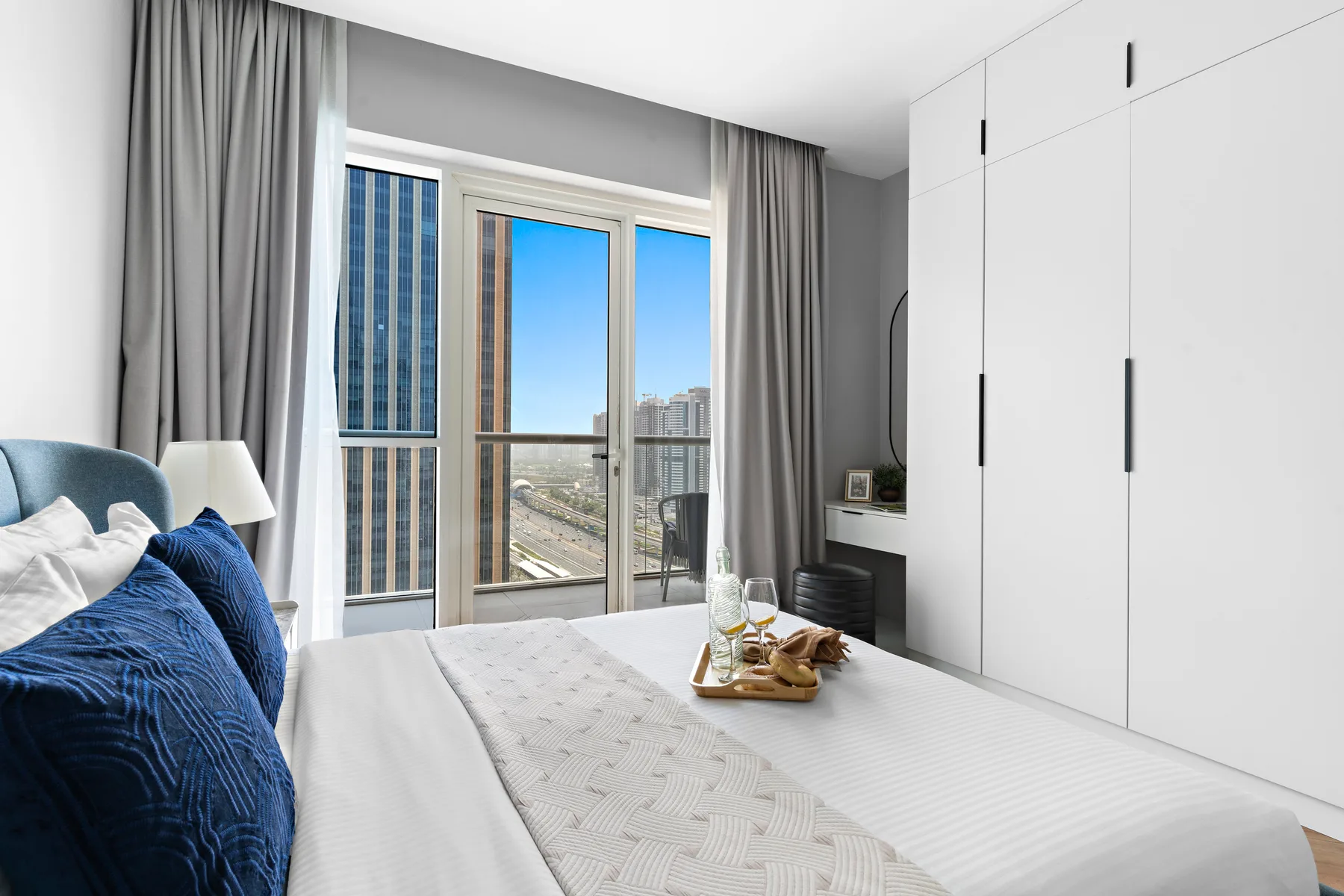 A bedroom with a neatly made bed beside a large window overlooking city buildings.