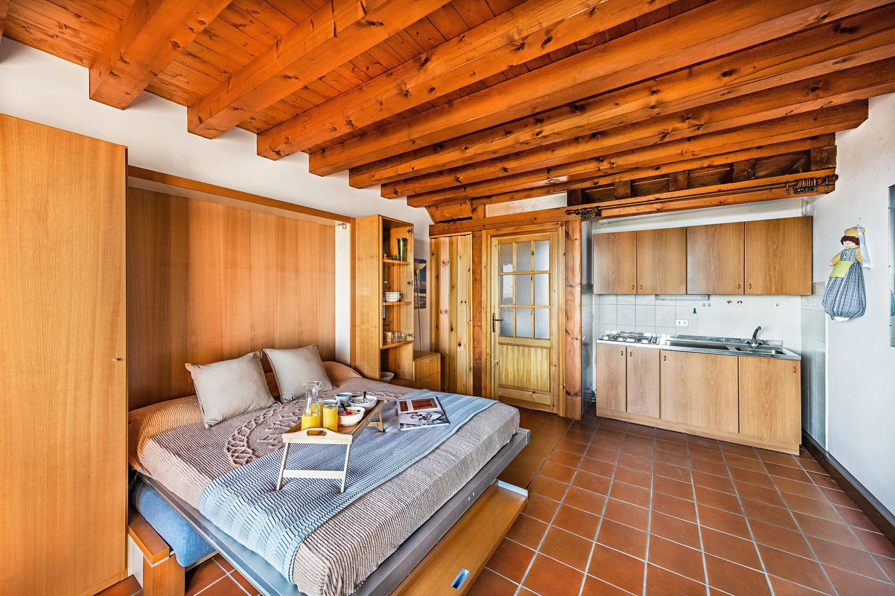 A bedroom with a bed beside a compact kitchen area and a wooden ceiling.